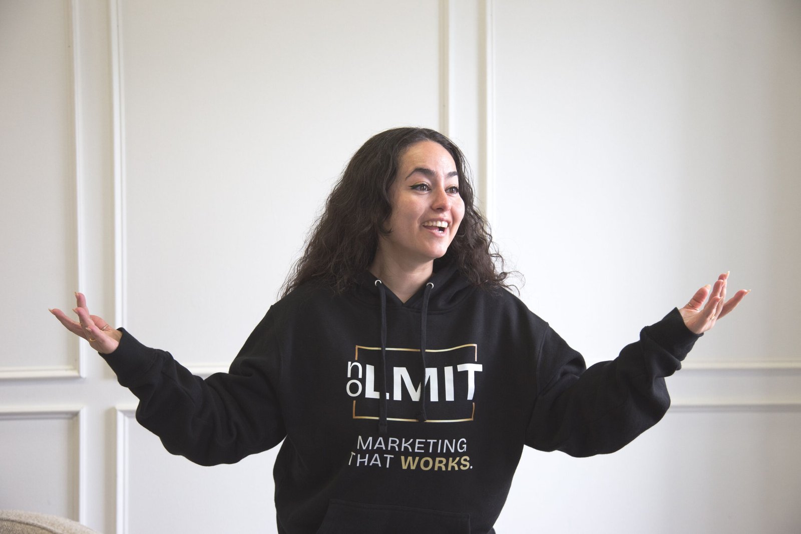 Smiling woman with curly hair wearing a black hoodie, arms extended outward in a bright room.